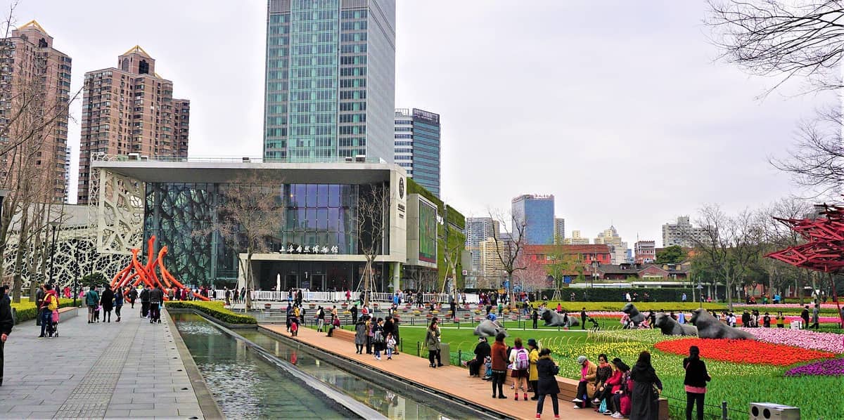 Eingang zum Shanghai Natural History Museum mit moderner Architektur, bunten Blumenbeeten, Skulpturengarten und Besuchergruppen an einem bewölkten Tag.