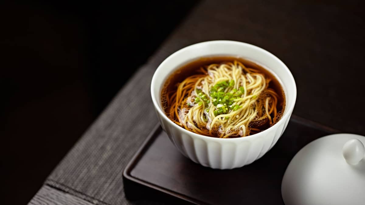 Eine große Schüssel Shanghai-Nudeln in kräftiger Brühe, garniert mit geschnittenen Frühlingszwiebeln, serviert auf einem dunklen Tablett mit einem Deckel daneben.