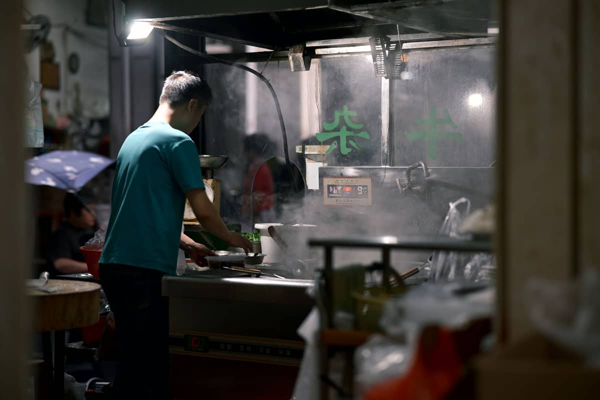 A man prepares noodles in a busy, steamy kitchen, likely in an Asian street food setting with visible kitchenware and atmospheric lighting.
