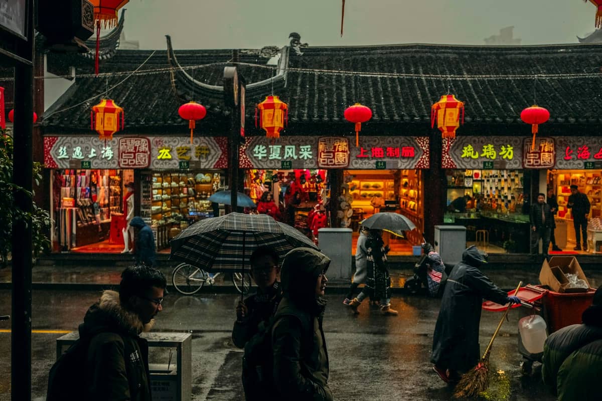 Rainy street scene in Shanghai with pedestrians under umbrellas in front of traditional lantern-lit storefronts.