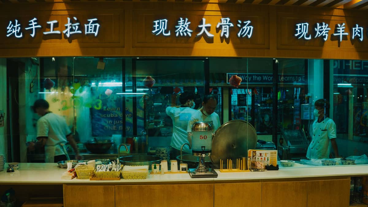 View through the glass of a noodle shop kitchen in China with chefs preparing different noodle dishes and a variety of ingredients on the counter.