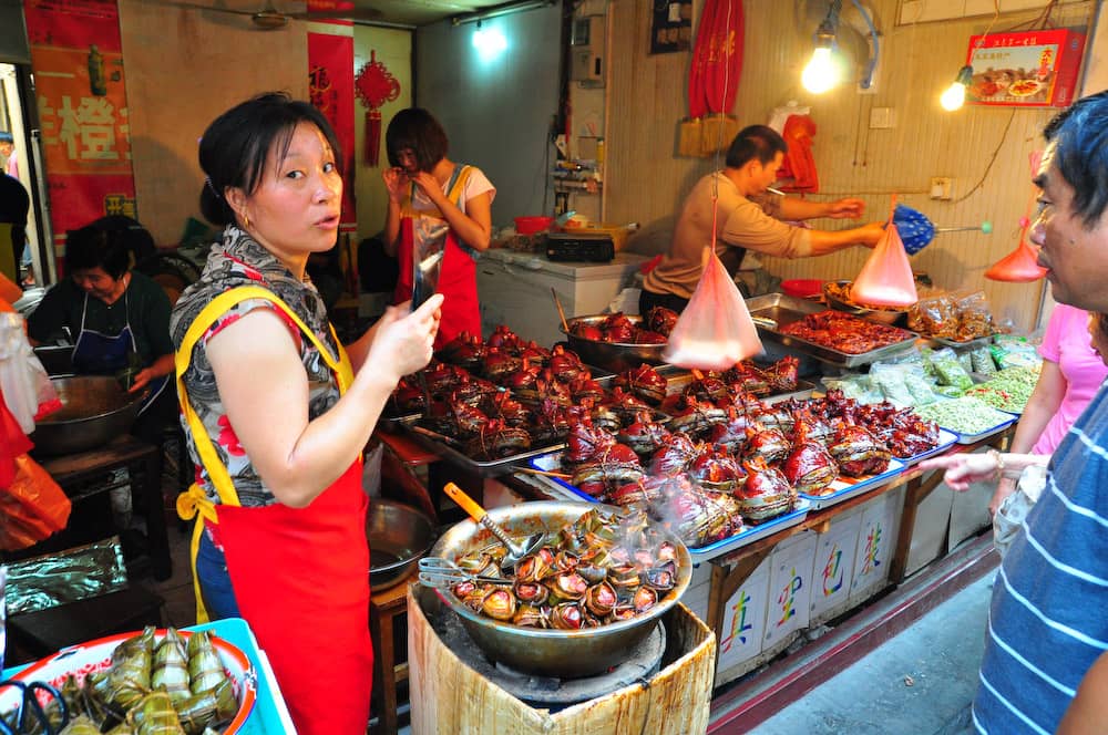 Un stand de street food animé à Shanghai vendant divers snacks traditionnels, avec des clients qui font leurs achats et un vendeur en tablier rouge qui prépare de la nourriture.