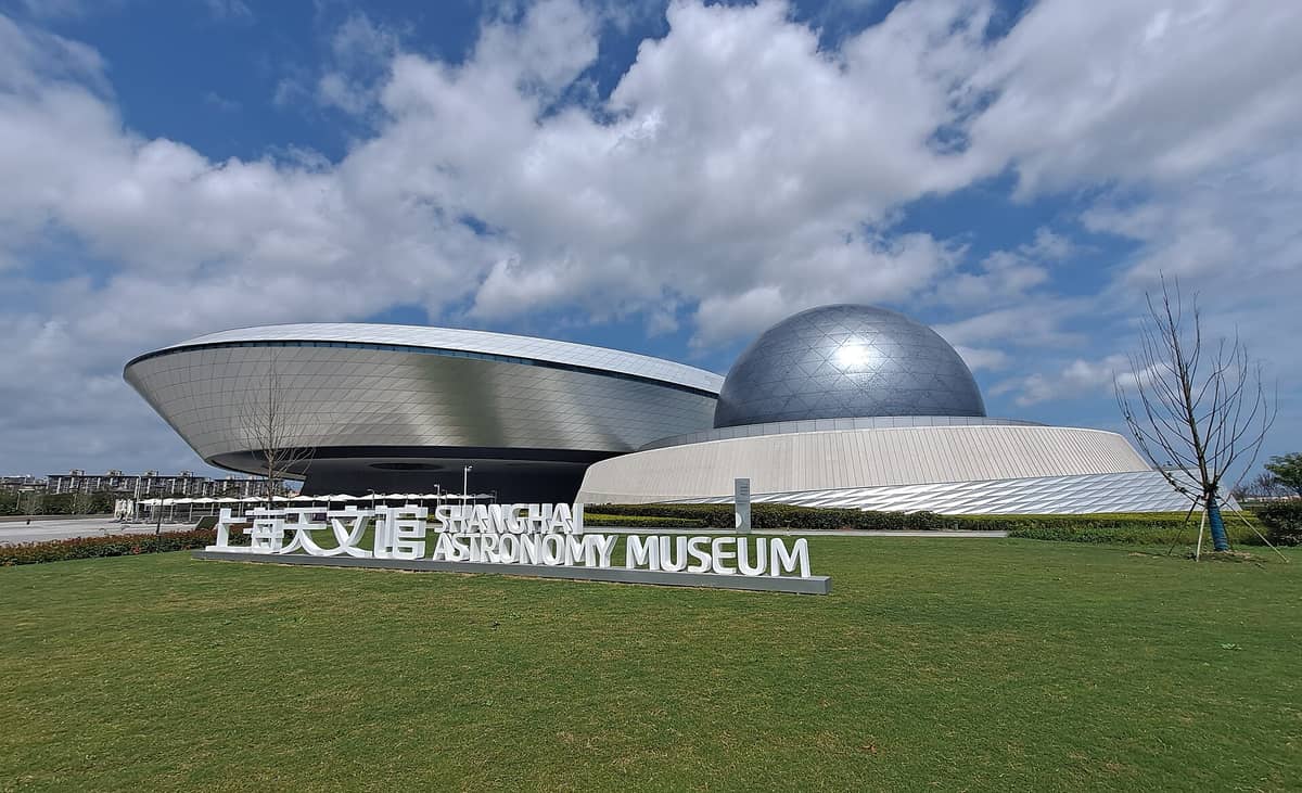 Weitwinkelansicht des Shanghai Astronomiemuseums mit seinem markanten geschwungenen Silberbau, der Kuppel und dem großen Museumsschriftzug vor blauem Himmel.