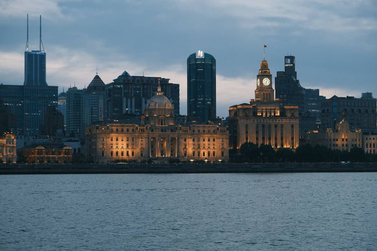 Dusk view of Shanghai Bund skyline across the Huangpu River featuring historic buildings and modern skyscrapers, with calm water in the foreground.