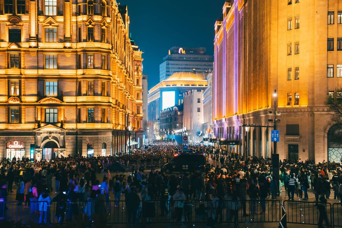 Large crowd of people at night along the illuminated Bund promenade in Shanghai, surrounded by historic buildings and vibrant city lights.