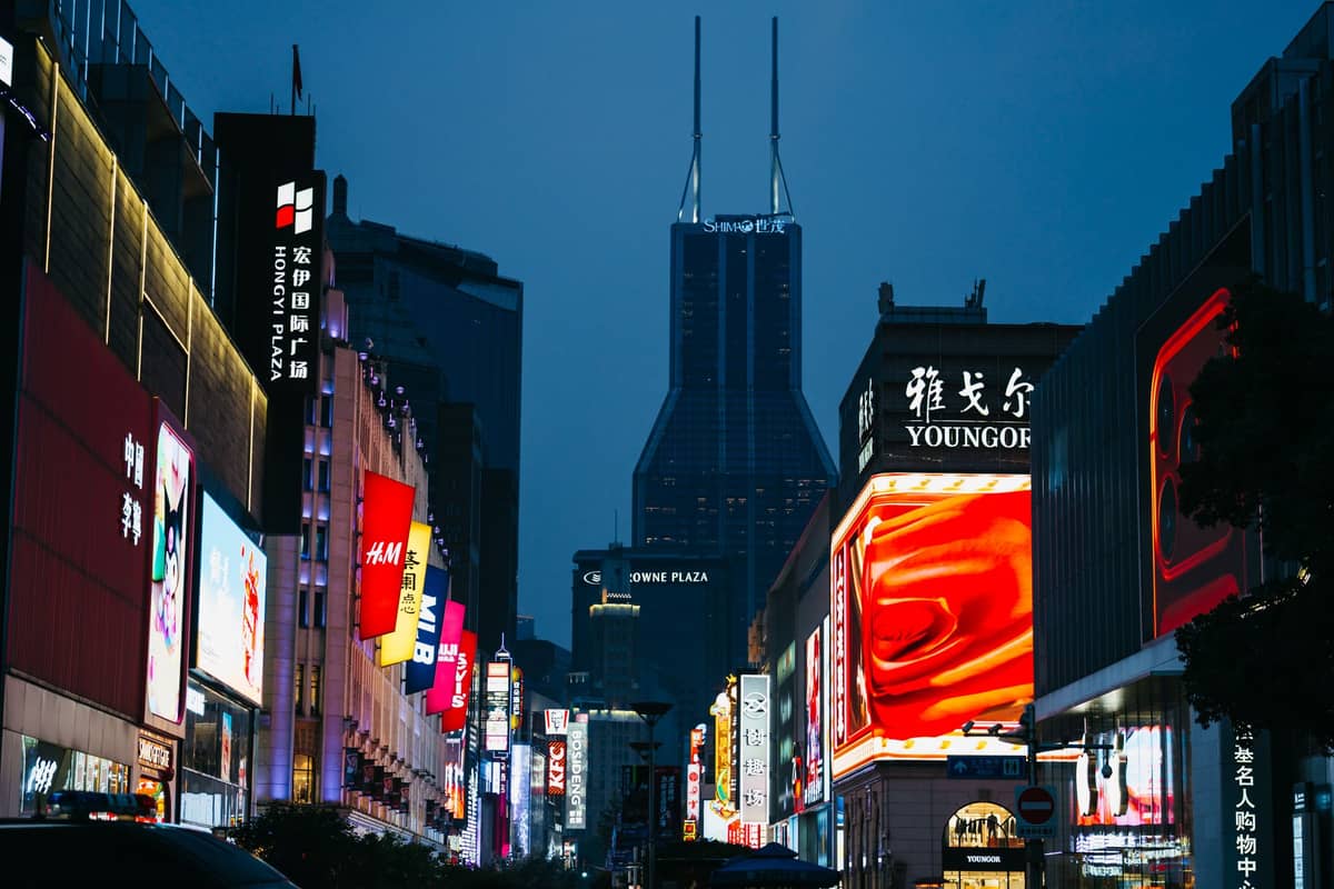 Nächtlicher Blick auf die Nanjing Road in Shanghai mit hell erleuchteten Geschäften, Werbetafeln und der markanten Krone des Tomorrow Square im Hintergrund.