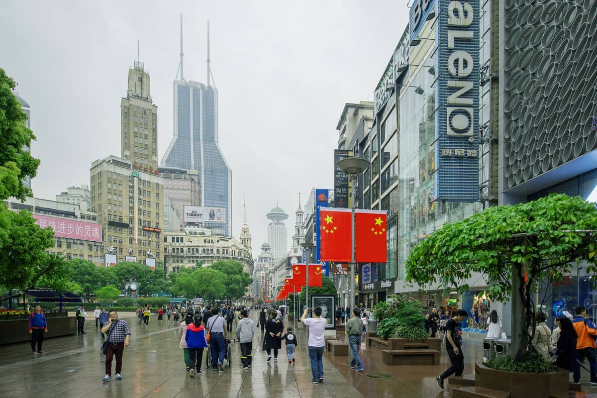 Wide view of Nanjing Road Pedestrian Street in Shanghai with crowds, modern buildings, and Chinese flags on a cloudy day.