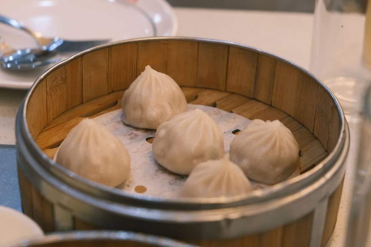 Nahaufnahme eines Bambus-Dampfgarkorbs mit vier Xiaolongbao-Suppen-Dumplings, Teller und Besteck im Hintergrund.