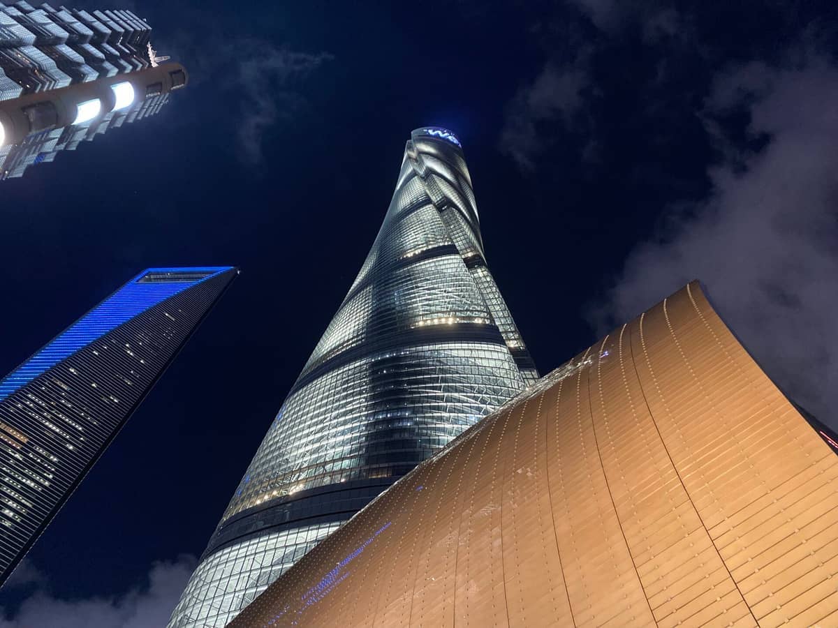 Nächtliche Aufnahme von unten auf den Shanghai Tower, flankiert von benachbarten Wolkenkratzern mit beleuchteten Fassaden und einem dramatischen bewölkten Himmel darüber.