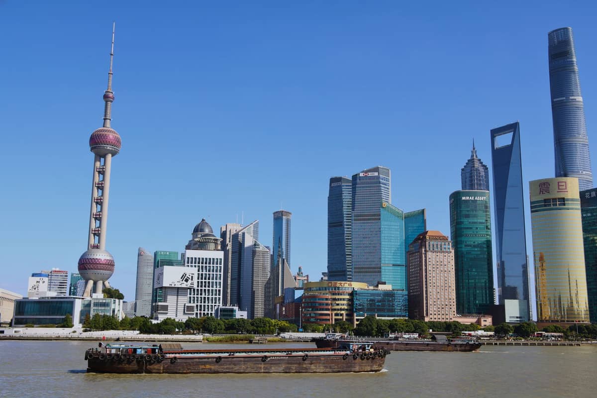 Tagsüber aufgenommene Skyline von Shanghai mit dem Oriental Pearl Tower, dem Shanghai World Financial Center und weiteren Wolkenkratzern am Huangpu-Fluss unter strahlend blauem Himmel.