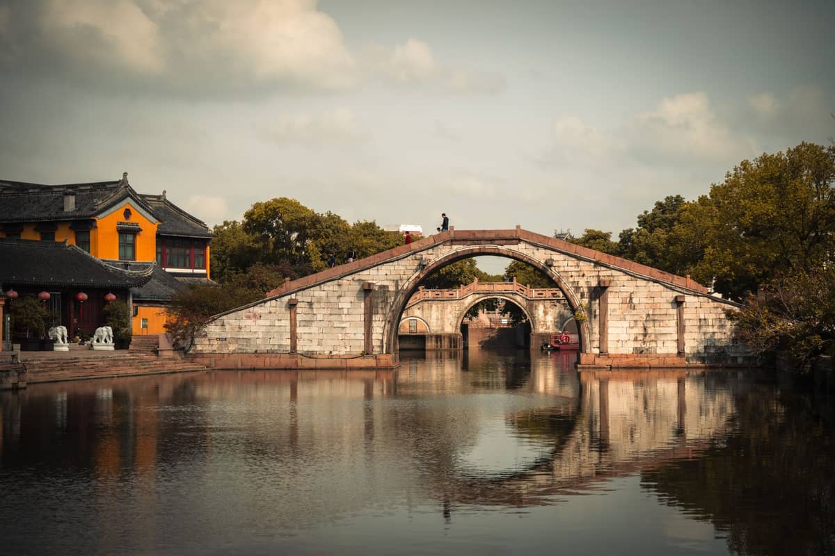 Arched stone bridge over canal with reflections, yellow building and trees, tranquil scene in an ancient Chinese water town.