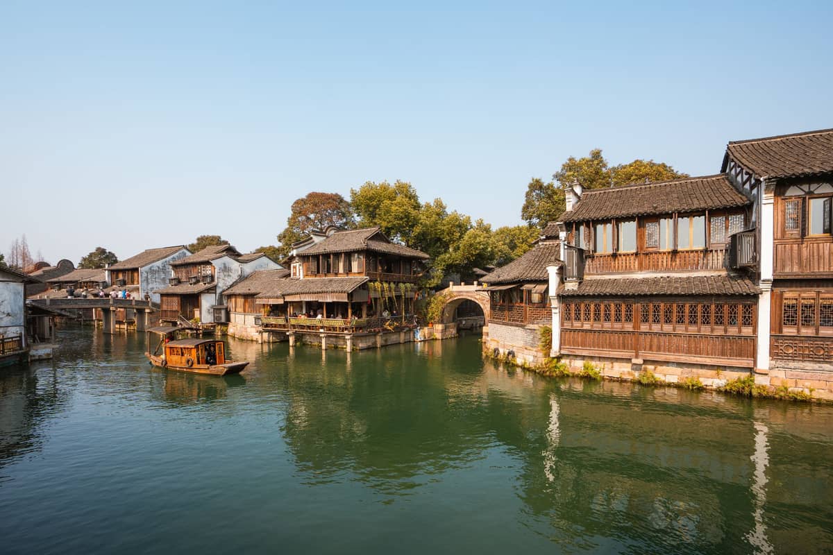 Scenic water town near Shanghai with traditional wooden buildings, arched stone bridge, and a small boat on a tranquil canal under clear blue sky.