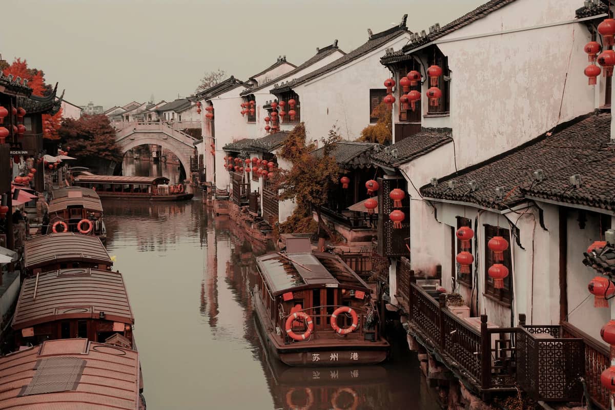 Traditioneller Kanal gesäumt von historischen weißen Gebäuden und roten Laternen in einer alten Wasserstadt bei Shanghai, mit Booten auf dem Wasser und einer gewölbten Steinbrücke im Hintergrund.