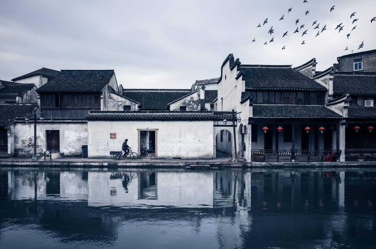 Traditional white-walled, black-roofed buildings line a canal in a water town at dusk, with red lanterns and reflections on the water.