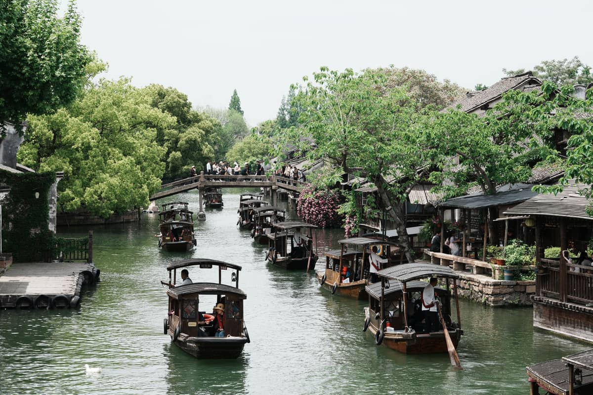 Traditional wooden boats glide along a canal flanked by old houses and willow trees, with a stone bridge ahead, in Wuzhen water town.