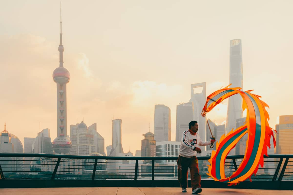 Ein Mann spielt mit einem bunten Drachenband vor der Skyline Shanghais im Sonnenuntergang, mit dem Oriental Pearl Tower und modernen Wolkenkratzern im Hintergrund.
