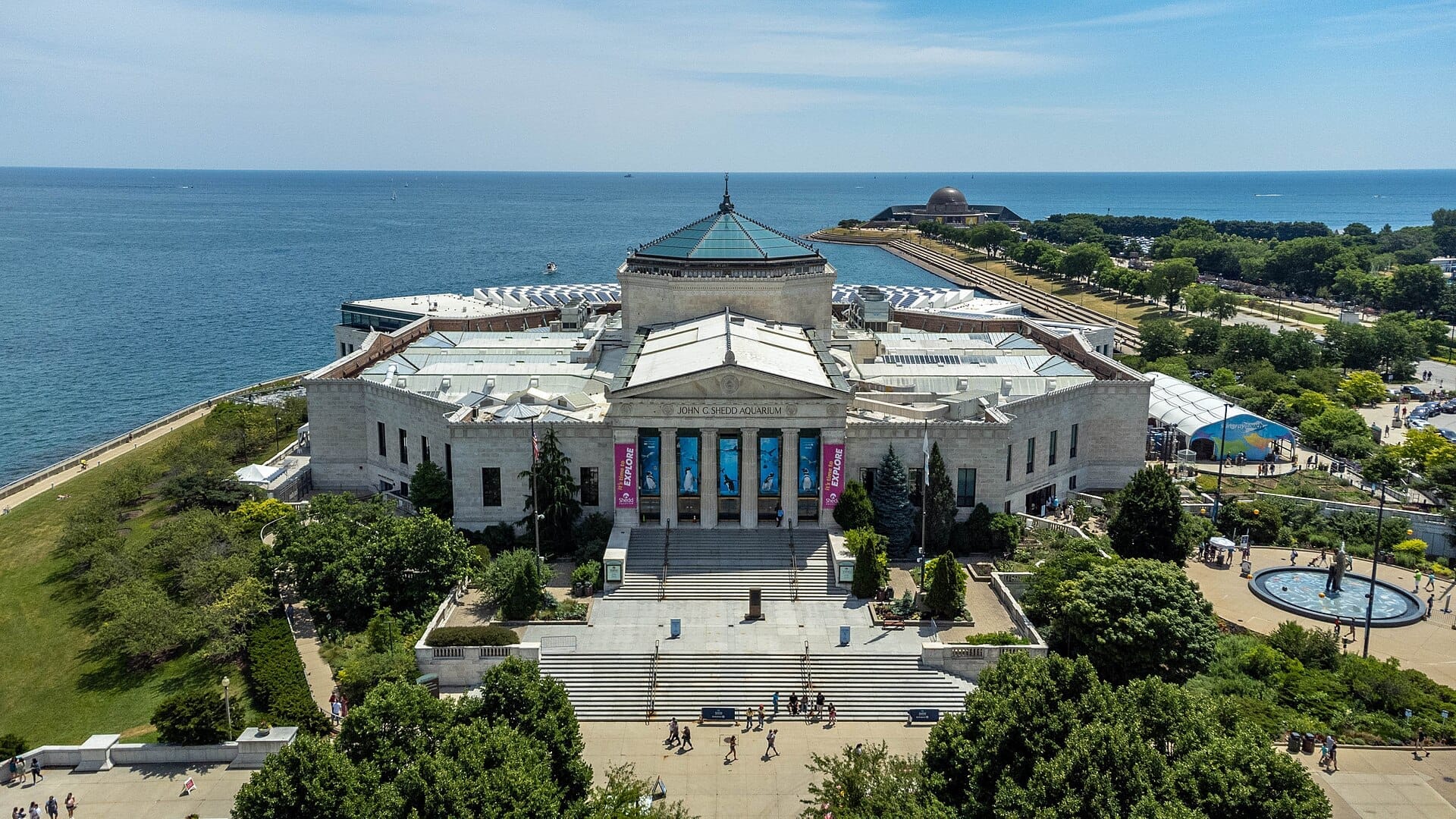 Vista aerea dello Shedd Aquarium a Chicago, con il suo grandioso ingresso, il verde circostante e il lago Michigan sullo sfondo in una giornata di sole.