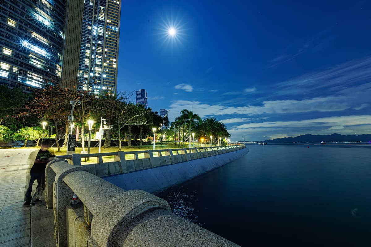 Vista noturna da orla de Shekou, em Shenzhen, com arranha-céus, palmeiras, luzes refletidas na água e montanhas ao fundo sob um céu enluarado.