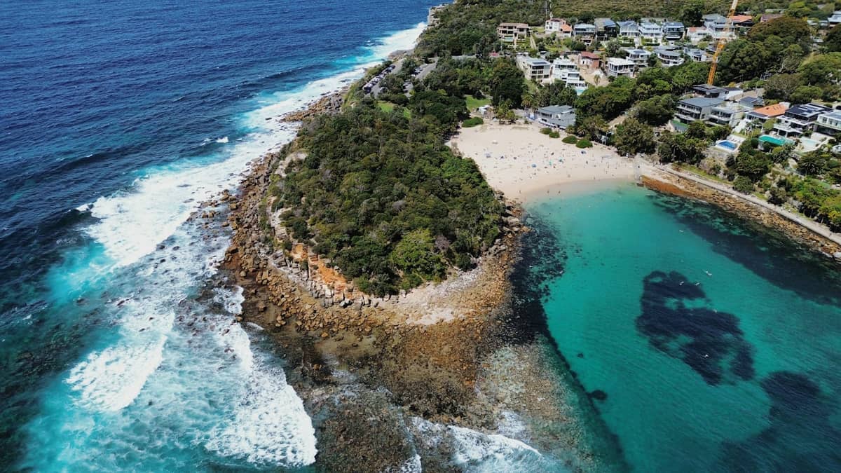 Vista aérea de Shelly Beach Manly con aguas turquesas cristalinas, costa rocosa, playa de arena y vegetación verde circundante con casas cercanas.