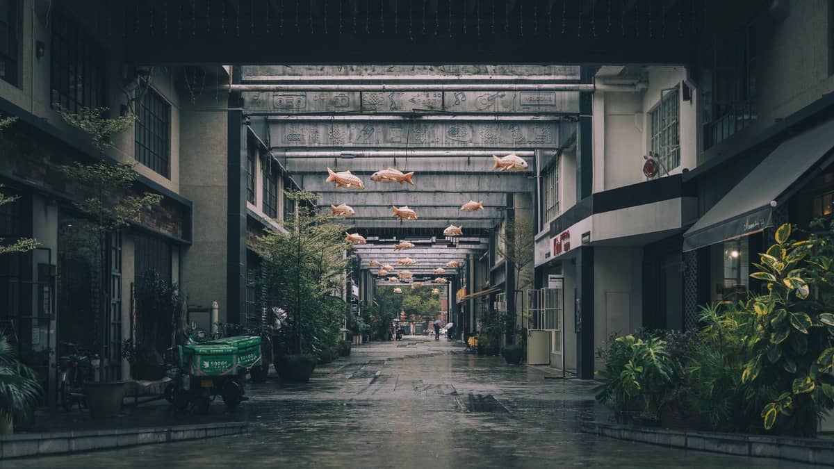 Um beco paisagístico de atmosfera sombria em uma área industrial renovada com esculturas suspensas, plantas e fachadas criativas, típico de distritos criativos como o OCT em Shenzhen.
