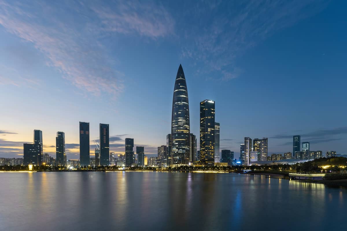 O moderno skyline de Shenzhen ao entardecer, com vários arranha-céus supertall, incluindo a icônica sede da China Resources refletida nas águas calmas da Baía de Shenzhen.