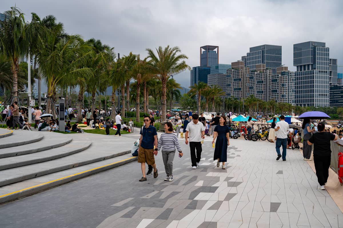 Multidão de pessoas passeando e relaxando ao longo de um moderno calçadão à beira-mar em Shenzhen, ladeado por palmeiras e arranha-céus ao fundo.