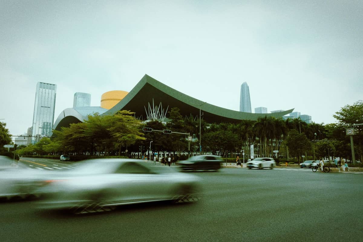 Vista ao nível da rua do Centro Cívico de Shenzhen, destacando seu telhado curvo e cúpula amarela, com carros passando em primeiro plano e arranha-céus ao fundo.