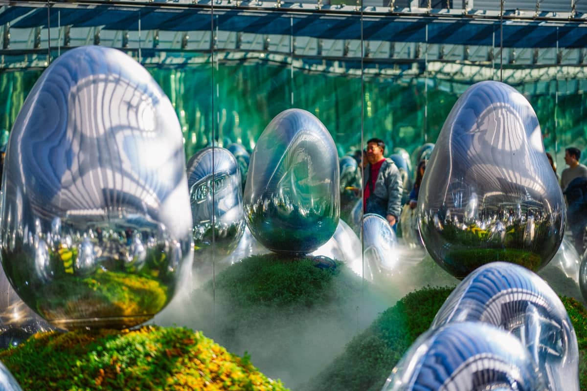 Esculturas reflexivas em forma de ovo e vegetação exuberante numa instalação high-tech com espelhos, com pessoas a observar a obra de arte.
