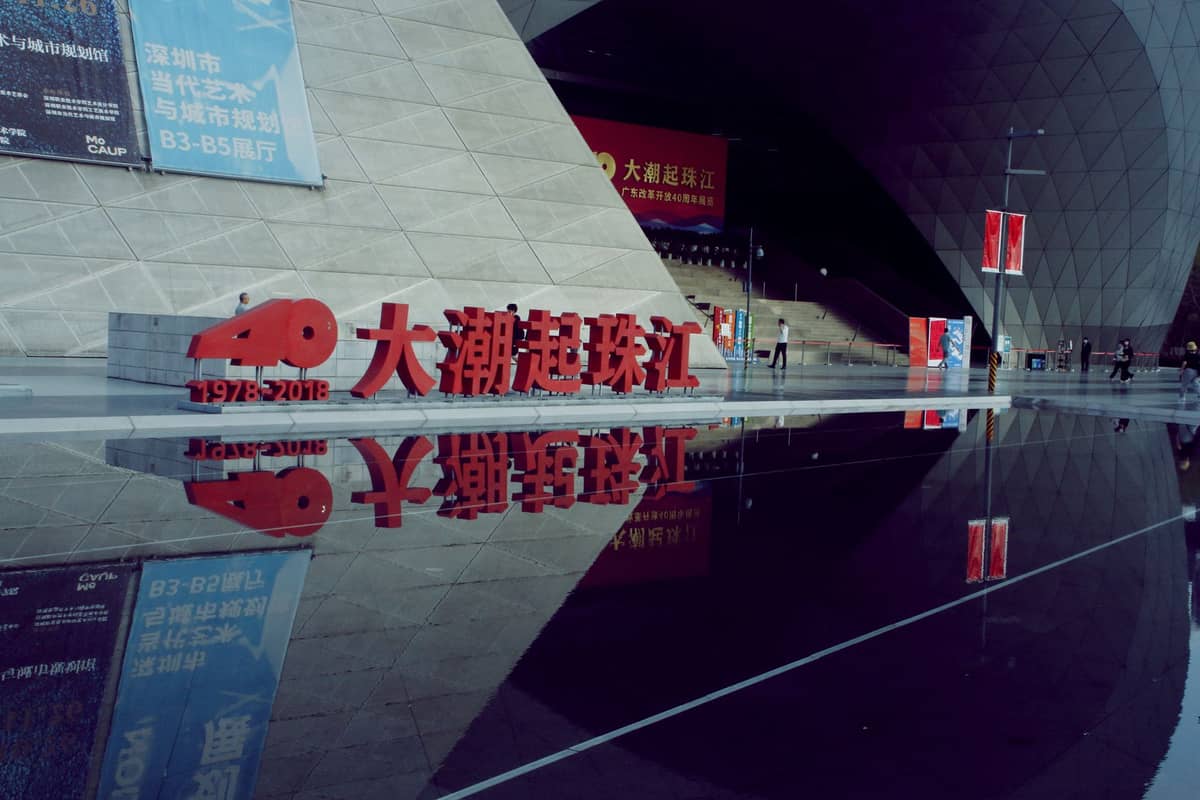 Fachada do imponente Museu de Arte Contemporânea e Planejamento Urbano de Shenzhen, com sua forma angular e moderna, entrada geométrica e reflexo espelhado numa praça molhada.
