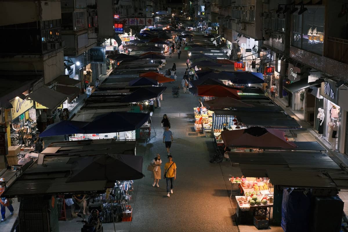 Vista de uma movimentada rua de mercado noturno ao ar livre com fileiras de barracas vendendo produtos e comida sob toldos coloridos, com pessoas caminhando entre elas.
