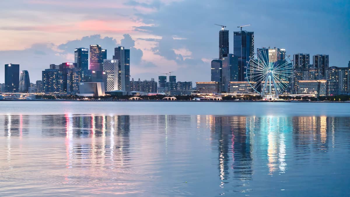 Horizonte moderno de Shenzhen com arranha-céus altos e uma grande roda-gigante iluminada à beira-mar ao entardecer.