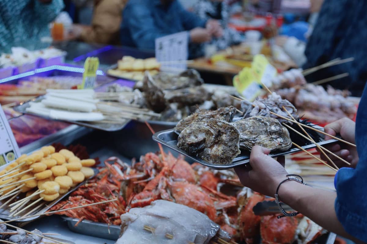 Pessoas selecionando frutos do mar e ostras em uma movimentada barraca de comida em um mercado noturno, com travessas de caranguejo e espetinhos visíveis
