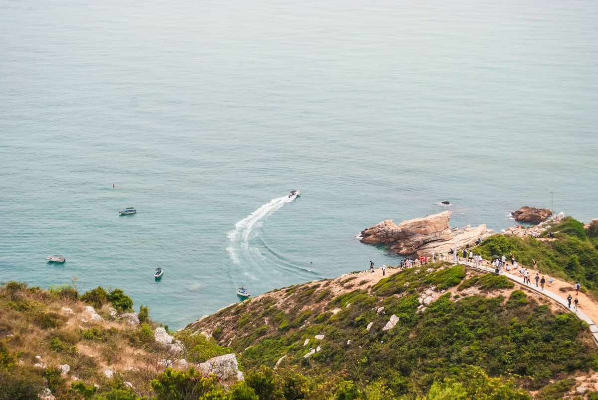 Uma trilha costeira panorâmica em Shenzhen com pessoas caminhando por um caminho dramático no alto de falésias com vista para o mar azul e a costa rochosa.