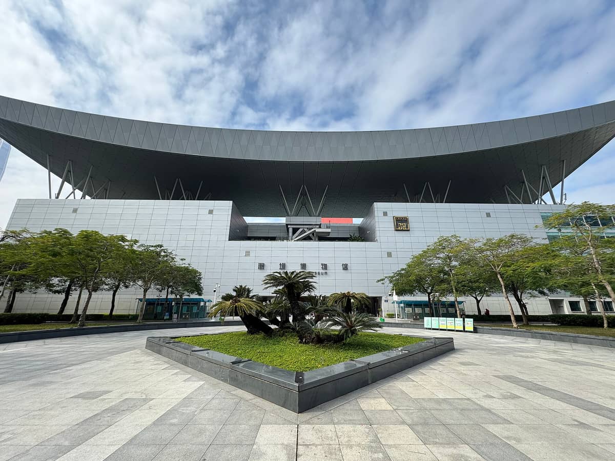 Vista ampla da entrada principal do Museu de Shenzhen com seu telhado moderno característico, a praça ao redor, árvores e céu azul límpido.