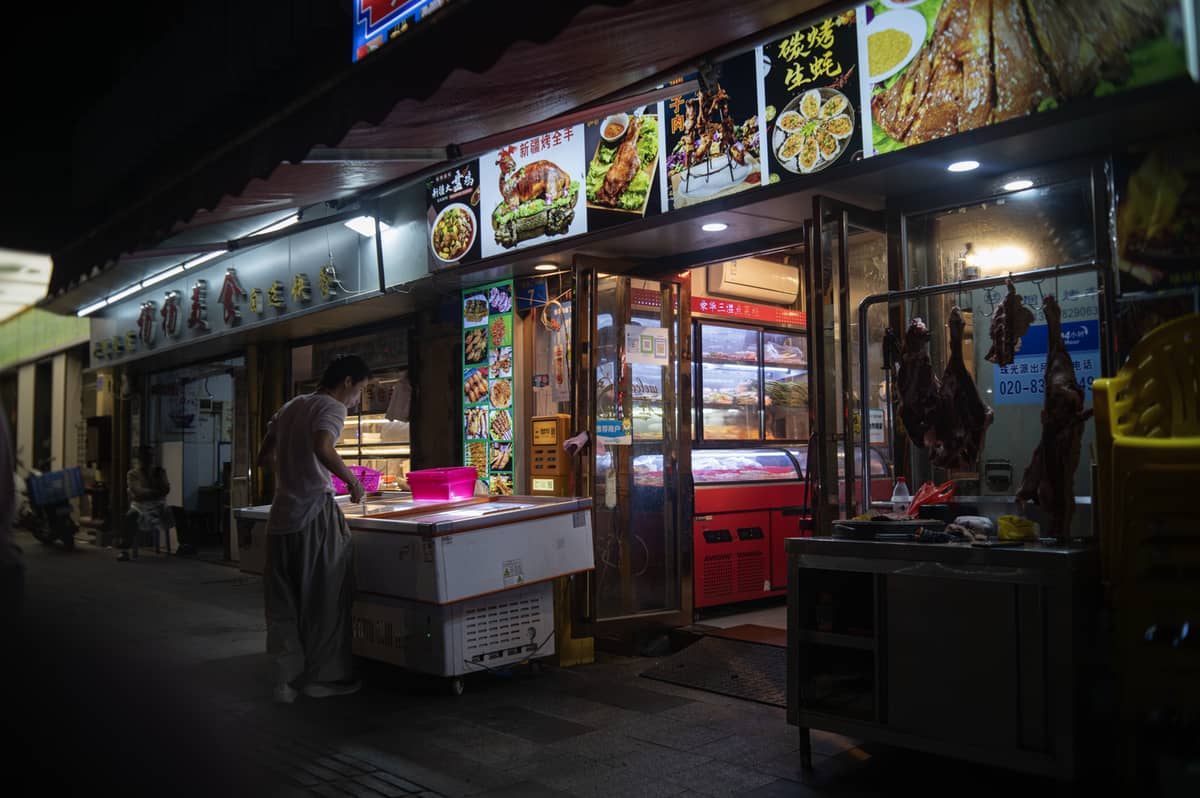Vista noturna de um vendedor de comida de rua chinesa preparando alimentos do lado de fora de uma loja bem iluminada com placas de comida e cardápios acima da entrada.