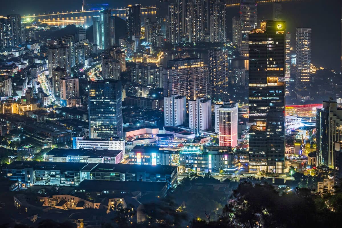 Vista noturna do distrito de Shekou em Shenzhen com a Sea World Plaza iluminada, apresentando arranha-céus, ruas movimentadas e área à beira-mar.