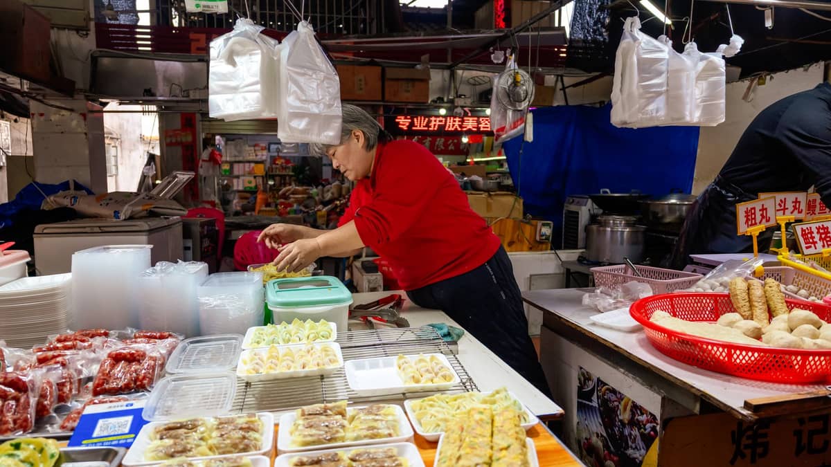 Barraca de comida em um mercado com um vendedor servindo pratos locais acessíveis em Shenzhen, rodeado de bandejas de comida preparada e placas coloridas.