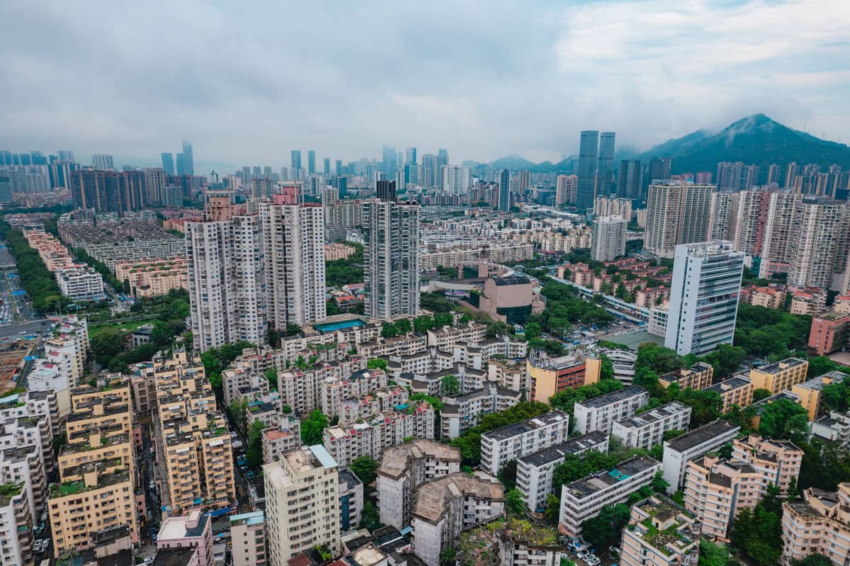 Vista ampla de um bairro densamente construído de Shenzhen com fileiras de edifícios de apartamentos de médio e grande porte e montanhas ao fundo.