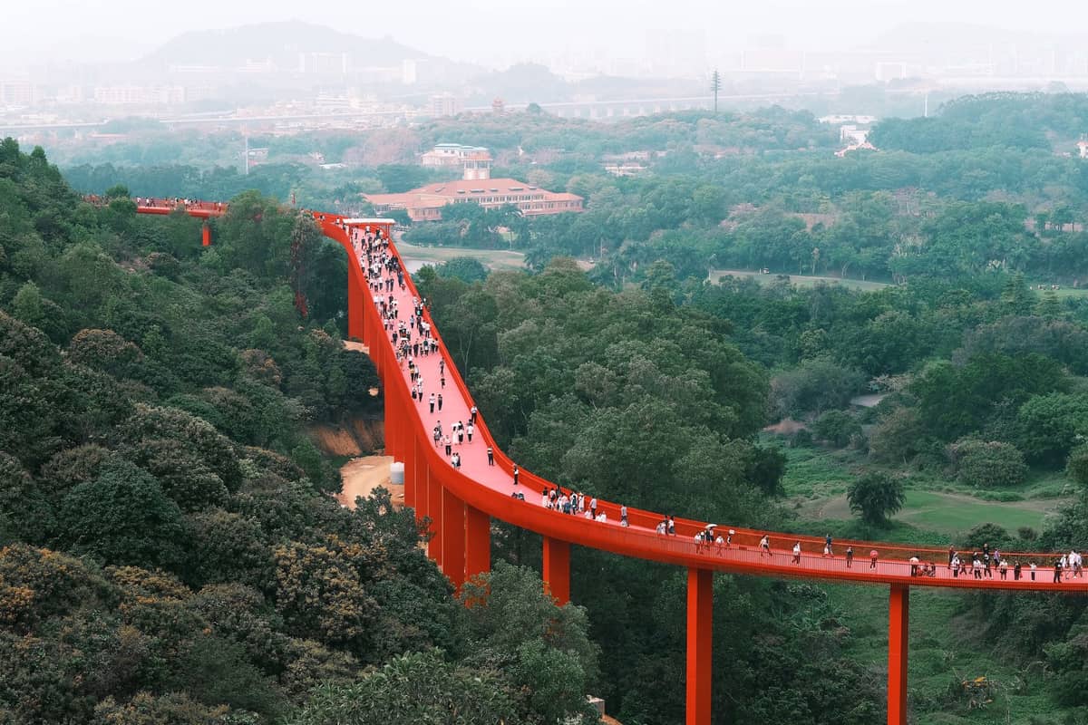 Passarela elevada vermelha com pessoas caminhando por colinas verdes, paisagem urbana e montanhas ao fundo em Shenzhen