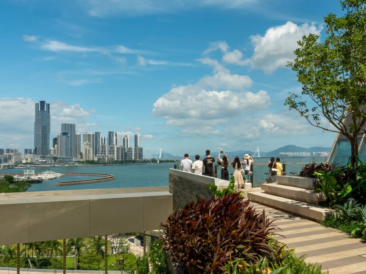 Pessoas desfrutando de uma vista panorâmica da via verde da orla de Shenzhen Bay, com arranha-céus da cidade, um exuberante parque ajardinado e céu azul ao fundo.