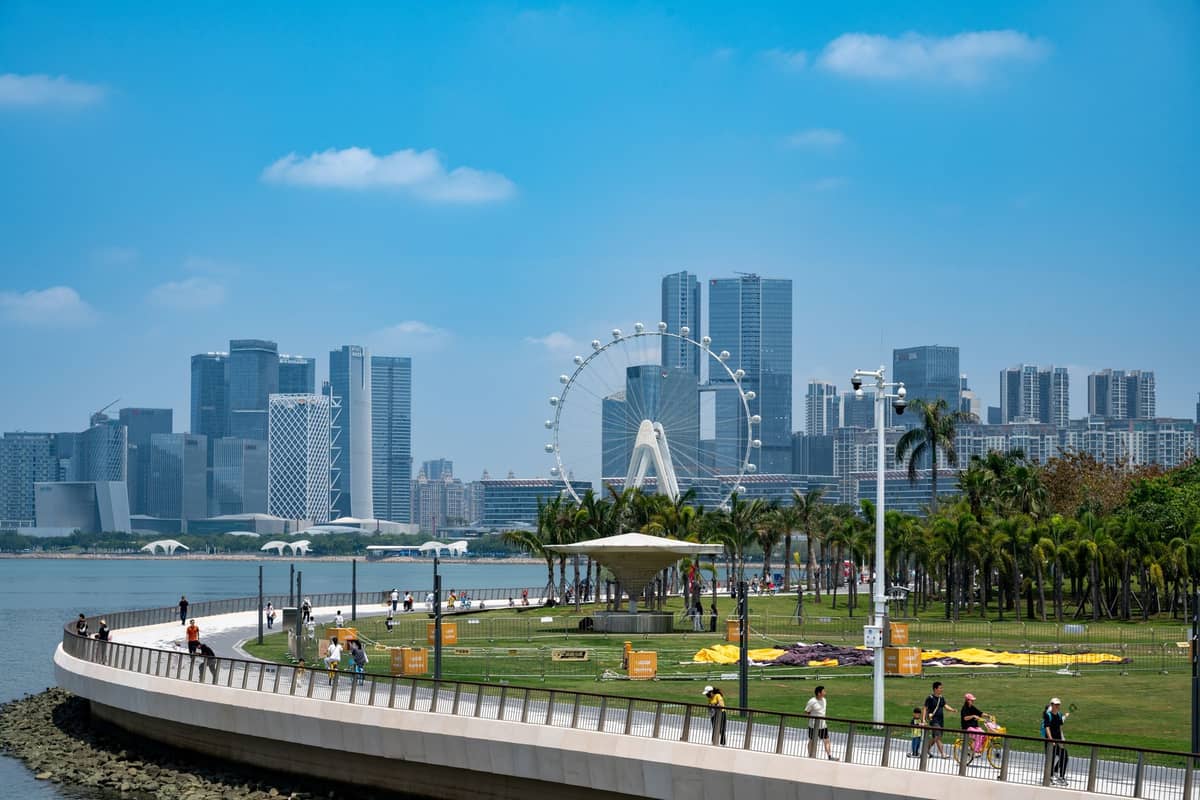 Vista panorâmica do Talent Park em Shenzhen, com gramado verde, palmeiras, pessoas caminhando e relaxando, e o horizonte da cidade com a icônica roda-gigante ao fundo.