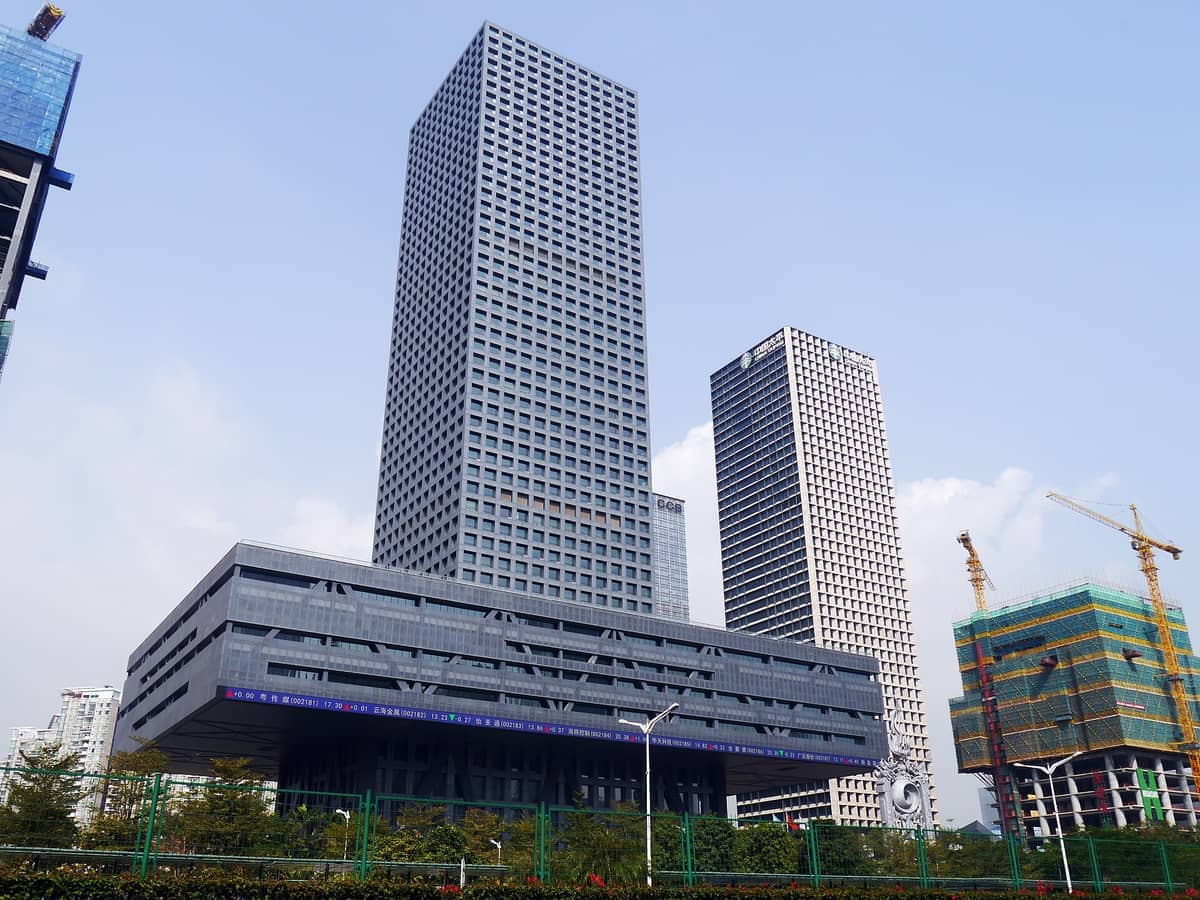 Vista frontal do Edifício da Bolsa de Valores de Shenzhen com seu design de pódio elevado, rodeado por arranha-céus modernos e guindastes de construção sob um céu azul claro.