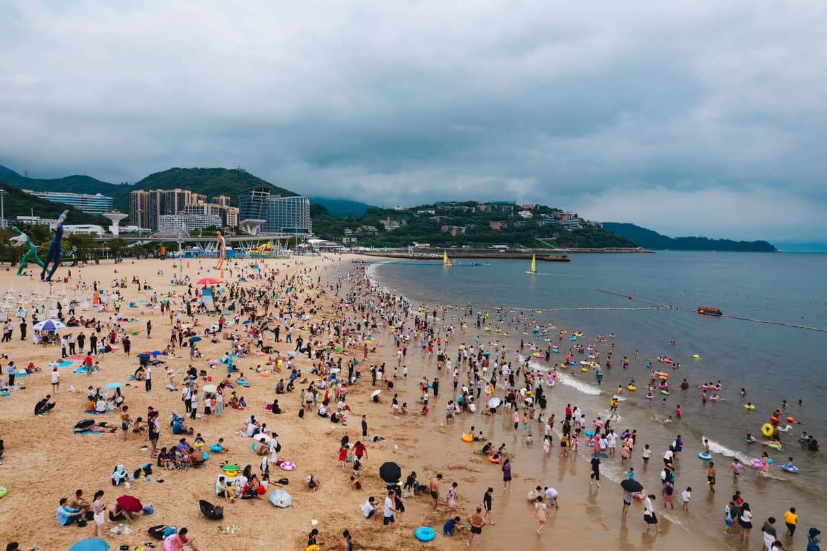Praia lotada em Shenzhen com muitas famílias e crianças aproveitando a areia e o mar, com colinas e edifícios ao fundo.
