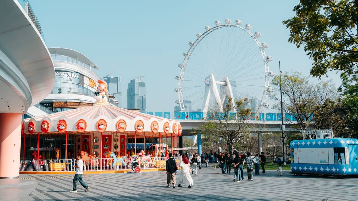 Área familiar na Baía de Shenzhen com um carrossel colorido, pessoas passeando, a grande roda-gigante da Baía de Shenzhen e os arranha-céus da cidade ao fundo.