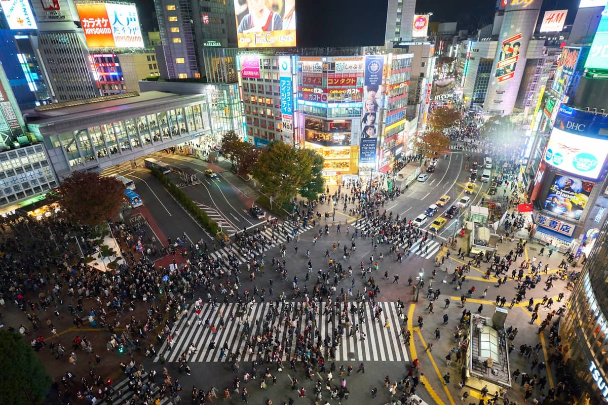 Aerial view of Shibuya Scramble Crossing at night, with large crowds crossing in multiple directions, surrounded by bright neon signs and busy city buildings.