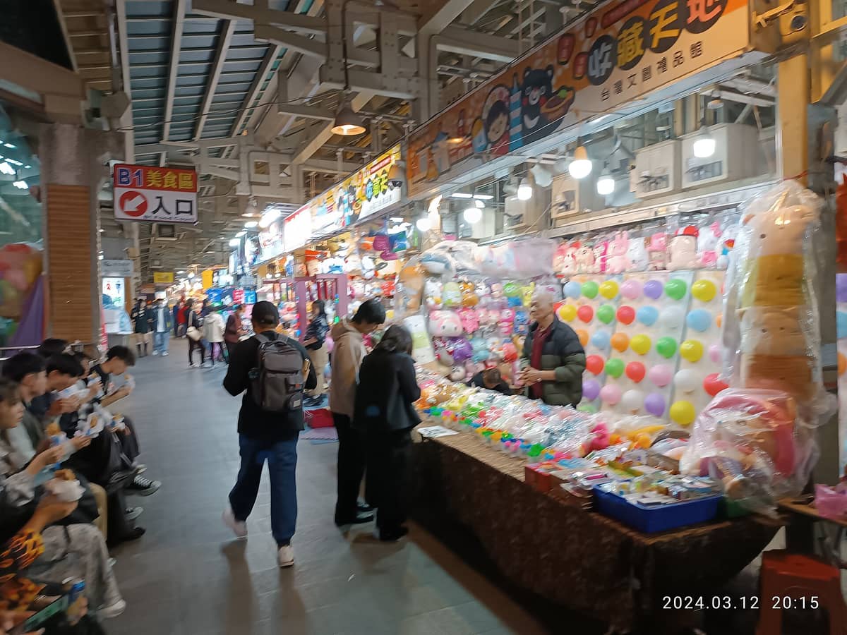 Bunte Innenstände mit Spielzeug, Luftballons und Snacks beim Shilin Nachtmarkt in Taipeh – abendliches Treiben und geschäftige Atmosphäre.