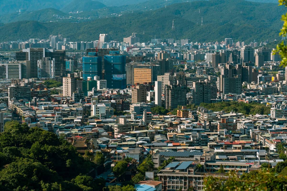 Panoramablick auf den Bezirk Shilin in Taipeh mit dicht bebauten Stadtvierteln, grünen Hügeln im Vordergrund und fernen Bergen unter blauem Himmel.