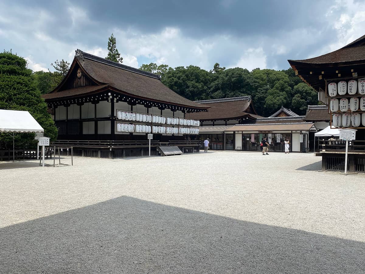 Wide view of the main courtyard and traditional wooden buildings of Shimogamo Shrine surrounded by forest, with white lanterns and a few visitors.