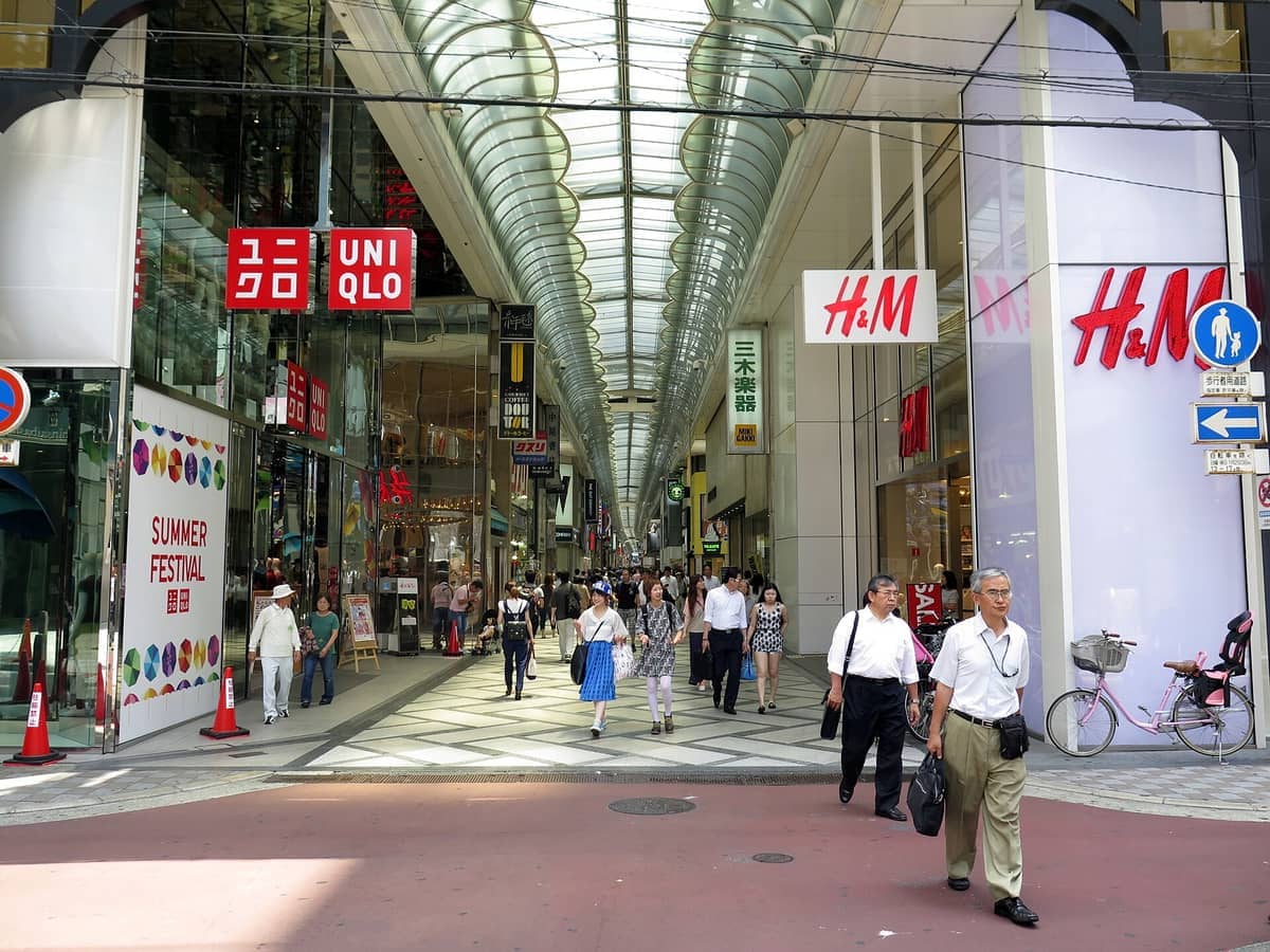 Shoppers walk through the covered Shinsaibashi arcade in Osaka, surrounded by modern storefronts like Uniqlo and H&M beneath a bright glass roof.