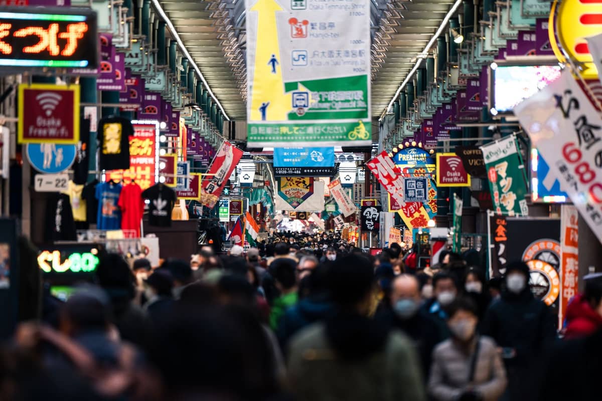 A bustling crowd walks through Shinsaibashi-suji Shopping Street, an indoor arcade with colorful signs, shops, and banners under a modern glass roof in Osaka.